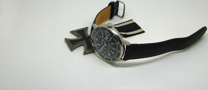 May include: A vintage wristwatch with a black leather band and a silver-colored case. The watch face is black with white and yellow markings. An Iron Cross medal and a black and white striped ribbon are also present.