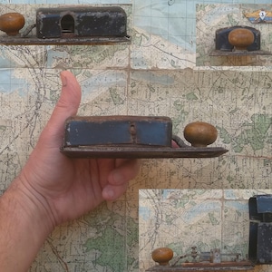May include: A vintage metal box with a wooden knob and a small opening on the top. The box is black with a brown wooden knob. The box is sitting on a map.
