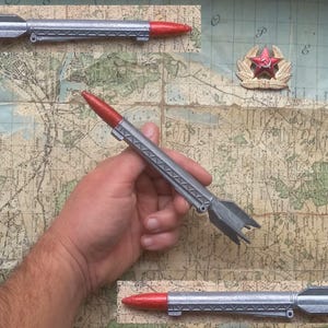Könnte beinhalten: Drei silberne und rote Spielzeugraketen mit einem roten Stern auf der Flosse. Die Raketen befinden sich auf einer Karte mit einem roten Stern und einem roten Stern mit einem Kranz.