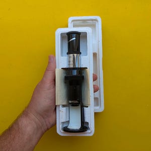 Puede incluir: Un objetivo de microscopio negro y plateado alojado en una funda protectora de poliestireno blanco moldeado. La lente cilíndrica presenta detalles metálicos y una base negra, asegurada dentro del embalaje de espuma a medida.
