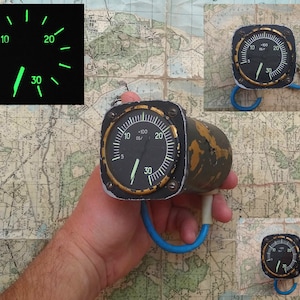 May include: A vintage aircraft instrument with a black face and a green needle. The instrument has a square shape and a gold-coloured bezel. The face of the instrument has markings for 0 to 30 and +100 deg/.