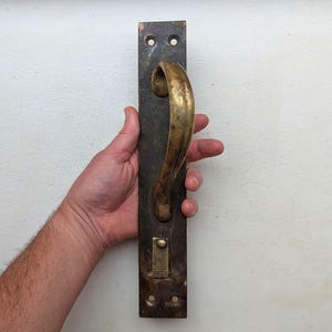 Può includere: Una maniglia antica con una piastra posteriore in metallo scuro e una maniglia curva color ottone. La maniglia è rettangolare con tre fori per viti nella parte superiore e inferiore. La maniglia è tenuta da una persona.