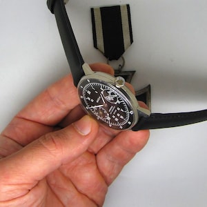 May include: A silver-cased wristwatch with a black dial and a black leather strap. The watch has multiple sub-dials and white numerals. A black and white ribbon is visible in the background.