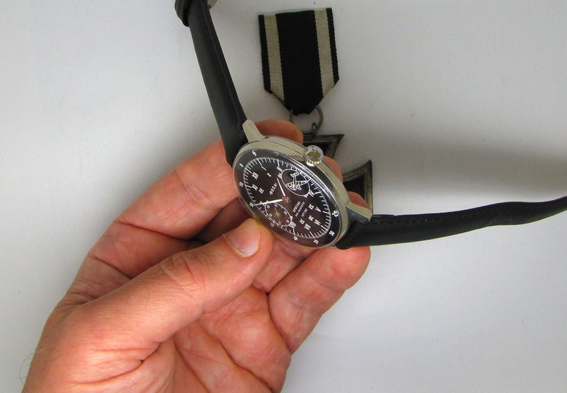 May include: A silver-cased wristwatch with a black dial and a black leather strap. The watch has multiple sub-dials and white numerals. A black and white ribbon is visible in the background.