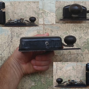 May include: A vintage black telegraph key with a round knob and a metal base. The key is being held in a hand, and there is a map in the background.