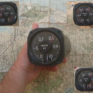 May include: A black analog gauge with white markings and a red needle, displaying numbers from 0.5 to 1.0. The gauge has the Russian word "число" and the letter "M" on its face. The gauge is held against a map background.