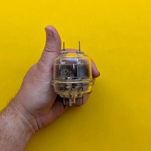 May include: A clear glass vacuum tube with internal metal components and two thin metal pins extending from the top. The cylindrical electronic component features visible markings including the text 65 and a logo on its transparent glass surface.