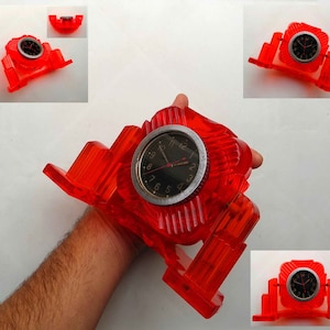 May include: A red plastic clock stand with a black clock face and silver hands. The clock is set on a stand that resembles a building with multiple levels.