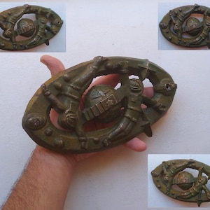 Puede incluir: Un adorno ovalado de metal de color bronce con un diseño detallado de dos figuras que sostienen una esfera. La esfera tiene un diseño en forma de estrella.