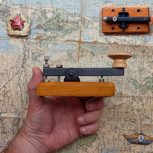 May include: A telegraph key featuring a rectangular wooden base, a black metal arm, and a light-colored wooden knob. The device includes adjustable metal screws and a pivot mechanism. A red star emblem and a small metal badge are visible nearby.