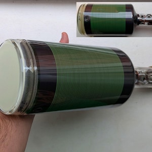 Puede incluir: Un tubo de vacío de vidrio transparente grande con un revestimiento rayado negro y verde. El tubo tiene una base de cerámica marrón con contactos metálicos.