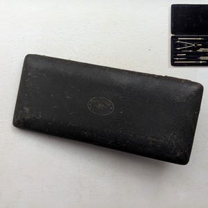 Könnte beinhalten: Ein schwarzes Lederetui mit einem Satz Zeichenwerkzeuge. Das rechteckige Etui hat ein kleines, ovales Emblem. Die silbernen Werkzeuge sind in einem mit schwarzem Samt ausgekleideten Fach angeordnet. Das Etui ist im Hauptbild geschlossen und im Inset geöffnet.