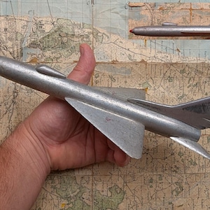 Può includere: Un modello di aereo color argento con un cono nasale rosso è tenuto in mano. L'aereo ha ali e una deriva. Una versione più piccola dell'aereo è sullo sfondo. Lo sfondo è una mappa vintage.