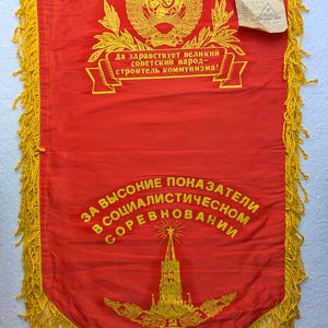 Op de afbeelding: Een rode vlag met gele franjes en geborduurde tekst in het Russisch. De vlag heeft een hamer-en-sikkel-embleem, een ster en een gebouw. De tekst luidt "Da zdravstvuyet velikniy sovetskiy narod - stroitel kommunizma!" en "Za vyssoniye pokazateli v sotsialisticheskom sorevnovanii".