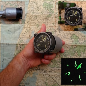 May include: A classic aircraft instrument with a black dial, displaying the numbers 1, 2, and 3, and the text "-1000 ОБОРТЫ". The instrument is held in a hand, with a map in the background. A second instrument and a starter motor are also visible.