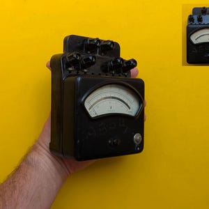 以下が含まれることがあります： カーブしたゲージと文字「AB04」が前面に表示された、ヴィンテージの黒い電気メーター。メーターの上部にはいくつかの黒いノブがあり、黄色の背景に対して保持されています。