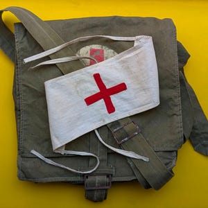 May include: An olive green canvas shoulder bag with a flap closure and a metal buckle, featuring a white fabric armband with a red cross symbol placed on top. The bag includes an adjustable shoulder strap and a faded circular emblem on the flap.