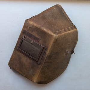 May include: A vintage brown leather welding helmet. The helmet features a rectangular viewing window with a dark lens and a hinged side closure. The leather shows signs of wear and age, with a slightly distressed appearance.