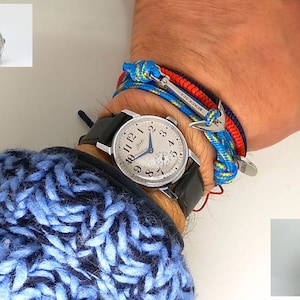 Puede incluir: Un reloj de pulsera plateado con una correa de cuero negro y una esfera azul. El reloj está en la muñeca de una persona con una pulsera de cuerda azul, roja y amarilla con un dije de ancla de plata.
