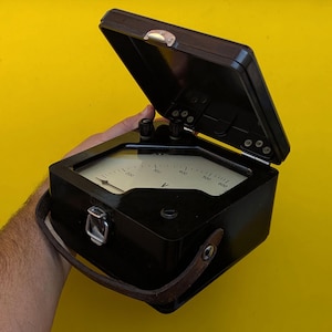 Op de afbeelding: Een zwarte analoge voltmeter in een rechthoekige behuizing met een scharnierend deksel. Het apparaat heeft een witte wijzerplaat met zwarte cijfermarkeringen en een naaldindicator. Een bruine leren draagriem is aan de zijkant bevestigd.