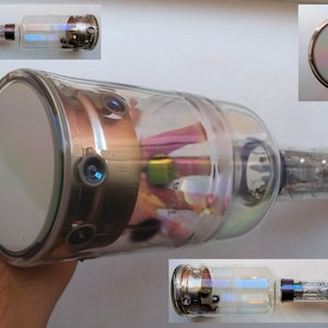 Könnte beinhalten: Eine große, durchsichtige Glas-Vakuumröhre mit einem silberfarbenen Metallsockel und einer kreisförmigen, irisierenden Glasscheibe an einem Ende. Die Röhre ist mit verschiedenen Komponenten gefüllt, darunter ein grüner und ein rosa Draht.