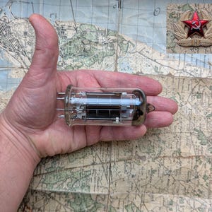 Puede incluir: Un tubo de vacío de vidrio transparente con una base de metal negro y una rejilla en el interior. El tubo mide aproximadamente 7,5 cm de largo y está siendo sostenido en la mano de una persona.