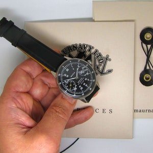 May include: A black-faced wristwatch with a black leather band is held in a hand. The watch face has white numerals and hands, with the brand name "DOXA" visible. The watch is set against a beige background with text and a nautical-themed logo.