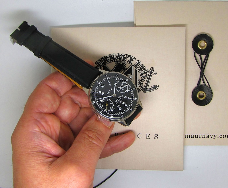 May include: A black-faced wristwatch with a black leather band is held in a hand. The watch face has white numerals and hands, with the brand name "DOXA" visible. The watch is set against a beige background with text and a nautical-themed logo.