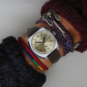 May include: A silver wristwatch with a white face and black hands. The watch has a brown leather band and is worn on a wrist with several bracelets, including a red, yellow, green, and blue bracelet, a navy blue rope bracelet with red and white accents, and a silver anchor bracelet with the text "Black Sea".
