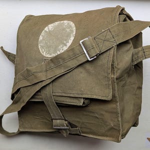 Könnte beinhalten: Olivgrüne Canvas-Tasche mit Schulterriemen und Metallschnallen. Die Tasche hat einen großen, verblassten weißen Kreis auf der Vorderseite. Ein rotes Sternemblem ist in der oberen linken Ecke sichtbar.