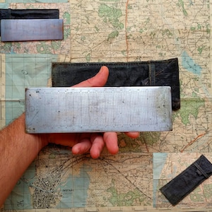 May include: A silver metal ruler with a grid pattern and numbers. The ruler is being held by a hand against a map background.
