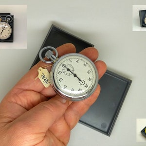 May include: A silver pocket watch stopwatch with a white face and black hands. The watch is in a black box with a tag that says "7474".