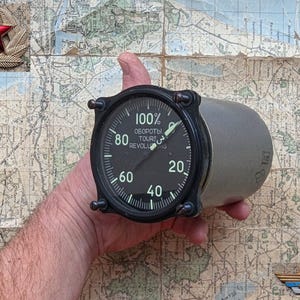 Puede incluir: Un instrumento de avión antiguo con una esfera negra, que muestra números y texto, incluyendo "100%" y "REVOLUTIONS". El medidor se sostiene contra un mapa antiguo. Un emblema de estrella roja es visible en la parte superior izquierda.