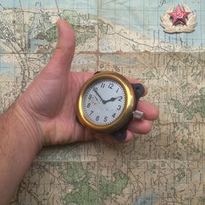 May include: A gold-coloured analogue clock with black hands and a white face. The clock has a black bezel and a small knob on the side. The clock is being held in a hand.