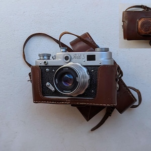 Puede incluir: Una cámara plateada y negra vintage con una funda y correa de cuero marrón. La cámara tiene una lente, diales y el texto "FED 3". La funda de cuero tiene una correa a juego y un emblema decorativo.