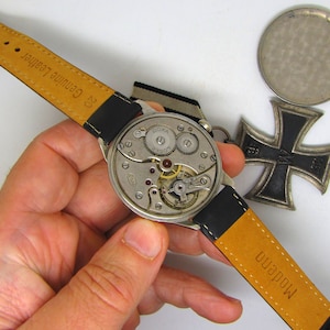 May include: A vintage DOXA watch with a black leather strap and exposed internal mechanisms. The watch is displayed with a silver-colored medal and a black and silver Iron Cross medal.