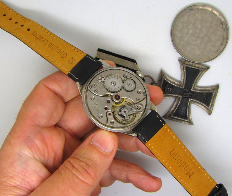 May include: A vintage DOXA watch with a black leather strap and exposed internal mechanisms. The watch is displayed with a silver-colored medal and a black and silver Iron Cross medal.