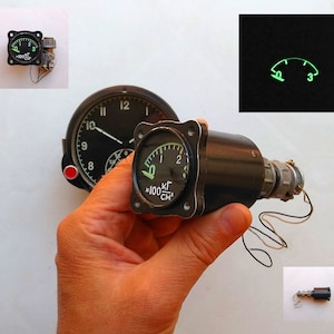 Może przedstawiać: Czarny i zielony analogowy wskaźnik z białą wskazówką i tekstem "x100 KG/CM2". Wskaźnik jest zamontowany na czarnej metalowej podstawie ze srebrnym pokrętłem i przymocowanym kablem.