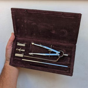 以下が含まれることがあります： 開いた状態の、ダークブラウンのベルベット裏地付きケースに入ったヴィンテージ製図セット。セットには、コンパス、ディバイダー、その他の製図ツールが含まれており、すべて銀色の金属製です。ケースは開いたままです。