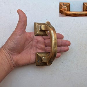 May include: A vintage brass drawer pull with a curved handle and square mounting plates. The hardware has a warm, aged patina, suggesting it's an antique or a reproduction. The pull measures approximately 13 cm in length.