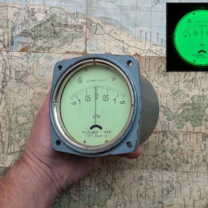Può includere: Un manometro blu chiaro con un quadrante circolare e un ago che punta allo zero. Il quadrante ha un'illuminazione verde e mostra numeri e segni bianchi. Il manometro è tenuto contro una mappa dettagliata.