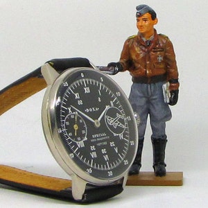 May include: A black-faced wristwatch with a silver case and a black leather band. The watch has white numerals and hands, with two smaller dials. A miniature figurine of a pilot stands nearby. The watch face reads "SPECIAL NON-MAGNETIC ANTITIME".