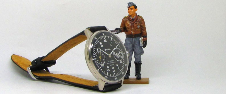May include: A black-faced wristwatch with a silver case and a black leather band. The watch has white numerals and hands, with two smaller dials. A miniature figurine of a pilot stands nearby. The watch face reads "SPECIAL NON-MAGNETIC ANTITIME".