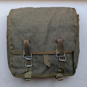 Puede incluir: Una bolsa cuadrada de lona verde oliva con una solapa frontal grande asegurada por dos correas tejidas marrones con hebillas de metal plateado. La bolsa tiene bordes reforzados y una construcción de tela texturizada con un diseño utilitario.