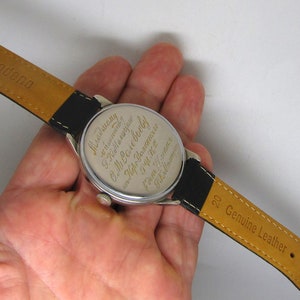 May include: A vintage wristwatch with a black leather band and a silver-colored watch face. The watch band is marked "Genuine Leather" and "Modena". The back of the watch face has engraved text.