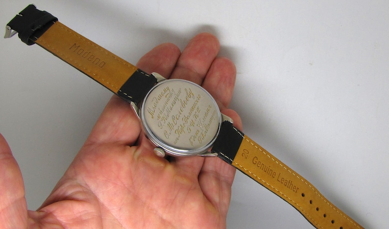 May include: A vintage wristwatch with a black leather band and a silver-colored watch face. The watch band is marked "Genuine Leather" and "Modena". The back of the watch face has engraved text.