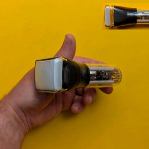 May include: A vacuum tube featuring a clear glass cylindrical body with internal metal components and a rectangular white ceramic top section. The base of the tube is tapered with a black coating, and the bottom end displays a circular array of pins.