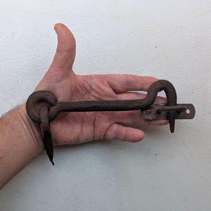 May include: A rusty metal hook latch with a curved handle and a flat plate. The hook is designed to be used as a door latch or gate latch.