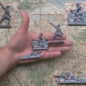 Puede incluir: Cinco soldados de juguete vintage hechos de metal. Los soldados están pintados en color plateado y representan a una variedad de personal militar, incluyendo un soldado lanzando una granada, un soldado conduciendo una motocicleta, un soldado disparando una ametralladora y un soldado de pie en posición de firmes.