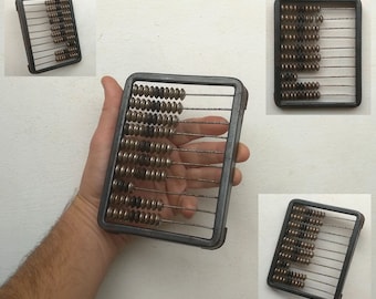 May include: A vintage metal abacus with brown beads on metal rods. The abacus is in a rectangular frame and has a hand holding it.